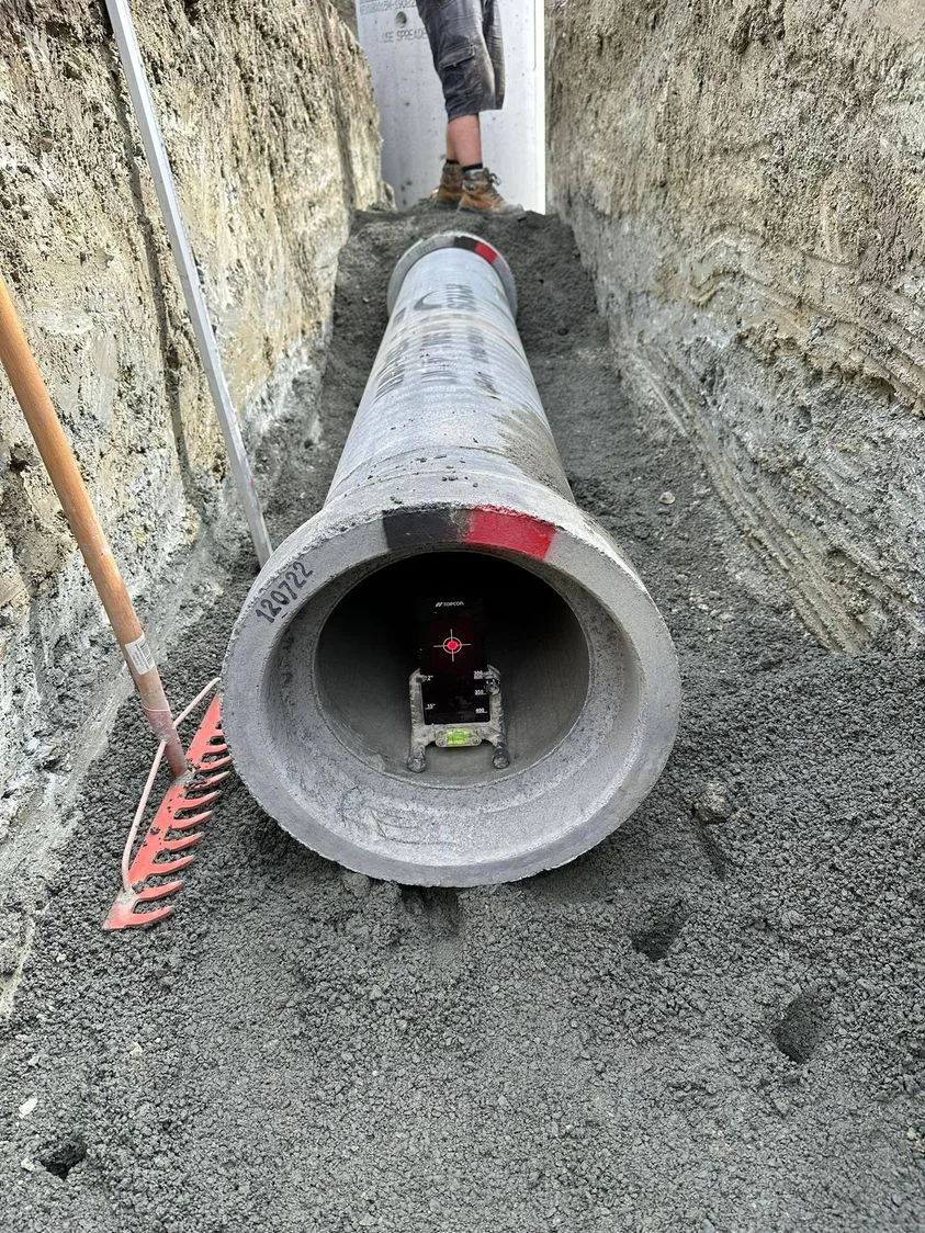New concrete drainage pipe laser-aligned for correct fall in an open trench on an Auckland site, gravel bedding visible underneath