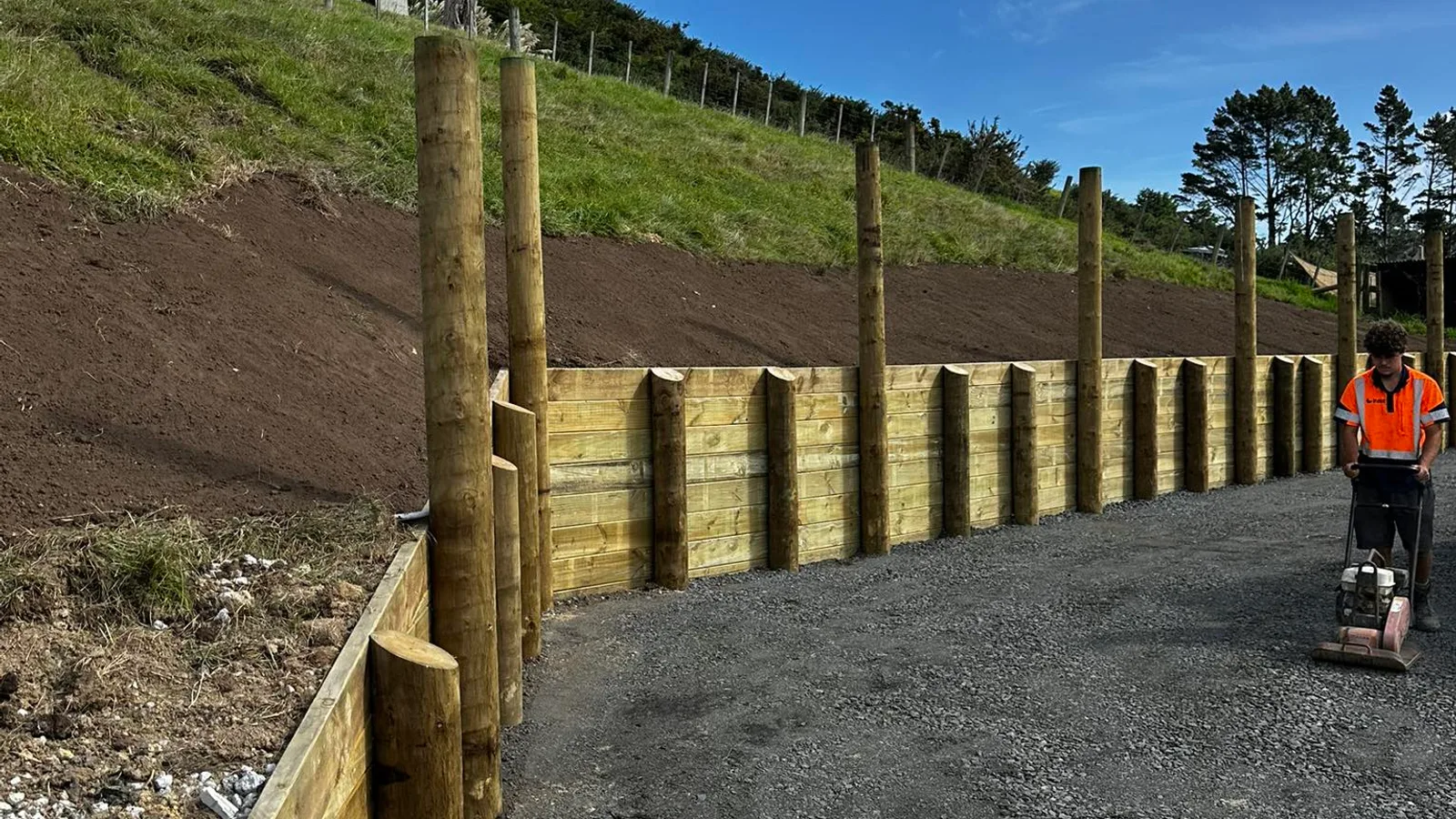 Timber retaining wall under construction on a sloped Auckland section with posts and rails visible, worker compacting gravel base